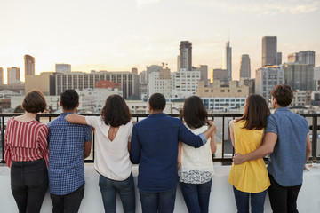Rear View Of Friends Gathered On Rooftop Terrace Looking Out Over City Skyline