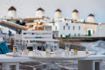 Leere Cocktail- und Schnaps-Gläser auf einem Tisch einer Bar vor den berühmten Windmühlen von Mykonos bei Sonnenaufgang, Kykladen, Griechenland