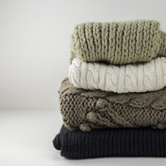 Warm woolen knitted winter and autumn clothes, folded in a pile on a white table. Sweaters, scarves. Place for text. Copyspace.