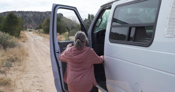 Adventurous senior woman stepping into large van and driving away on dirt trail