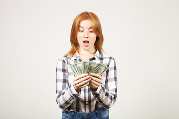 Young beautiful redhead woman screaming in rejoice, laughing w/ open mouth, fistful of one hundred dollar bills like fan. Excited attractive female with lots of cash. Background, copy space, close up.