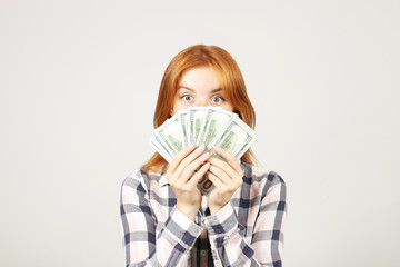 Young beautiful redhead woman covering her face with fistful of money holding bunch of one hundred dollar bills like fan. Excited attractive female with lots of cash. Background, copy space, close up.