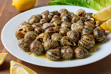 Tasty fried snails with lemon in white plate on brown wooden background.