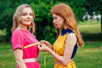 ginger redhead irish norwegian girl dressmaker seamstress measures the body with a measuring tape of beautiful and young blonde woman in pink dress in summer park. tailor and customer on outdoors