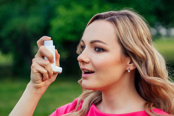 fighting asthma and bronchitis. active cheerful beautiful young blonde woman in a pink dress using the inhaler in the summer park . concept of spring allergy