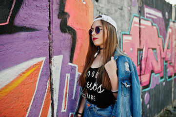 Stylish casual hipster girl in cap, sunglasses and jeans wear, listening music from headphones of mobile phone against large graffiti wall.