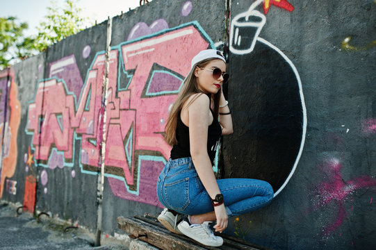 Back of buttocks stylish casual hipster girl in cap, sunglasses and jeans wear, listening music from headphones of mobile phone against large graffiti wall with large tnt bomb.