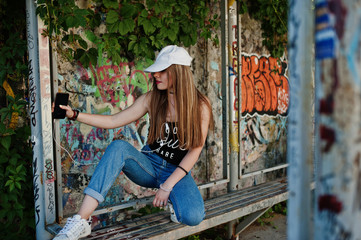 Stylish casual hipster girl in cap and jeans wear, listening music from headphones of mobile phone against large graffiti wall.