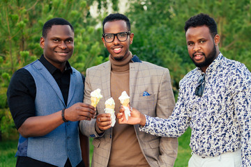 a group of three black men in stylish suits a meeting in a summer park. African-Americans friends hispanic businessman eating vanilla white sweet ice cream in a waffle horn picnic outdoors