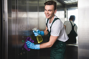 Young cleaning company worker cleaning elevator and smiling at camera