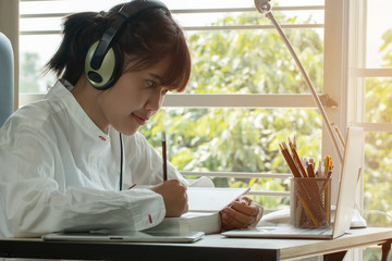 Student learning online study concept: Beautiful Asian girl listening with Headphones and laptop, sitting smile writing notes in textbook at her desk in home for e-learning in educational technology