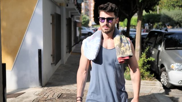 Young handsome man strolling, taking a walk along Italian town street in summer day, with beach towel over his shoulders