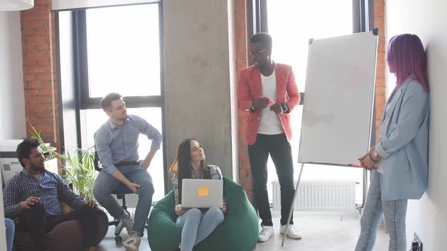 Young African American businessman in stylish red jacket giving a presentation to his colleagues at work as they plan a new business strategy together in the modern office meeting room.