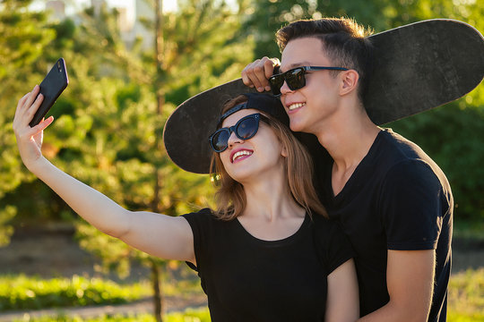 active couple in love hugging in the summer park. beautiful woman in a cap and sunglasses is holding a skateboard(longboard) and photographed take pictures selfie on phone with man . guy and girl date