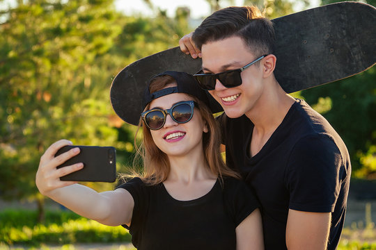 active couple in love hugging in the summer park. beautiful woman in a cap and sunglasses is holding a skateboard(longboard) and photographed take pictures selfie on phone with man . guy and girl date