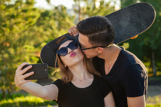 active couple in love hugging in the summer park . beautiful woman in a cap and sunglasses is holding a skateboard( longboard) and photographed take pictures selfie on phone with man . guy kisses girl