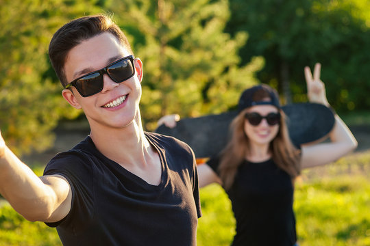 cool guy and a lovely girl in sunglasses, jeans and a black T-shirt skateboarding holding hands photographed take pictures selfie on phone in the summer park
