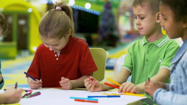 Medium shot of group of adorable diverse children of elementary school age talking to each other when drawing picture using colored felt tip pens