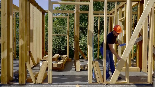 adult man is making measurements of wooden beams in a frame of new house under construction, using tape and level