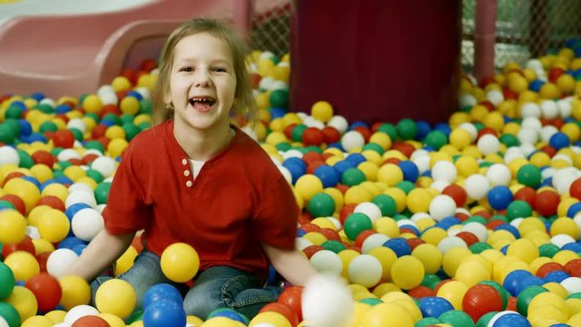 Medium shot of joyful little girl smiling at camera and throwing colorful hollow plastic balls when having fun in ball pit in children play center