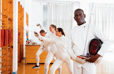 Active african american male fencer in uniform standing with mask and foil at fencing room