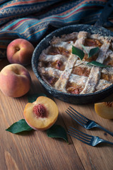 Peach pie in a cast-iron frying pan lies on a wooden table. On the table lie cut peaches, green leaves, fabric, knife and forks.