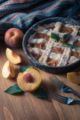 Peach pie in a cast-iron frying pan lies on a wooden table. On the table lie cut peaches, green leaves, fabric, knife and forks.