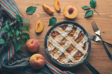 Peach pie in a cast-iron frying pan lies on a wooden table. On the table lie cut peaches, green leaves, fabric, knife and forks.
