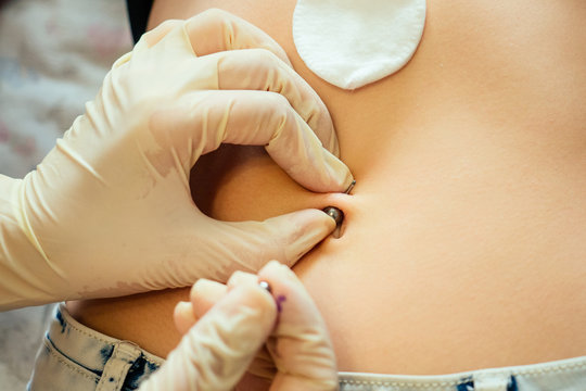 hand in a rubber glove close-up of master prepares to pierce the navel by belly of a young woman with a bandage and cotton on her stomach. process navel ring piercing