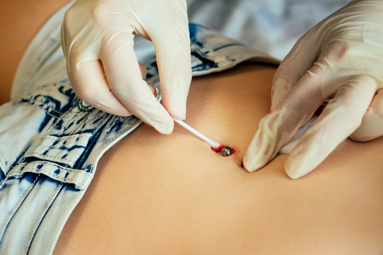 hand in a rubber glove close-up of master pierce the navel by belly of a young woman with a bandage and cotton on her stomach titanium piercing . process navel ring piercing