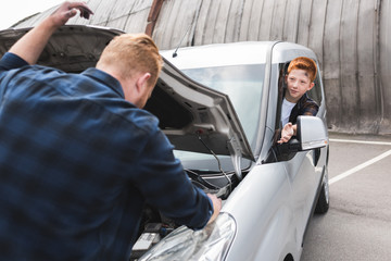 father repairing car with open hood, son sitting at driver seat