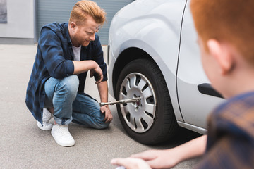 father changing tire in car with wheel wrench, son sitting near