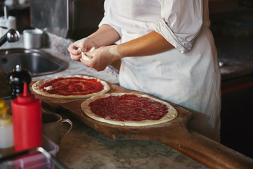cropped shot of chef putting cheese pieces onto pizza at restaurant kitchen