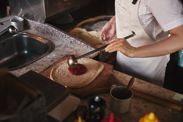 cropped shot of cook pouring ketchup onto pizza dough at restaurant kitchen