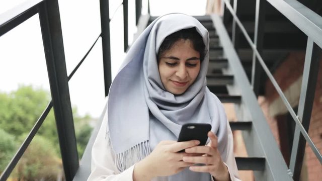 Attractive young woman with black hair wearing traditional muslim clothes smiling, sitting on iron stairs in a city and texting. Tilt down real time establishing shot