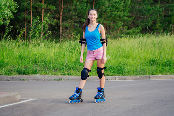 attractive young athletic slim brunette woman in pink shorts and blue top with protection elbow pads and knee pads on roller skates in the park