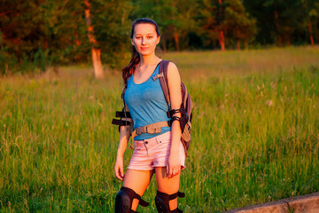 attractive young athletic slim brunette woman in pink shorts and blue top with protection elbow pads and knee pads on roller skates in the park in the evening at sunset