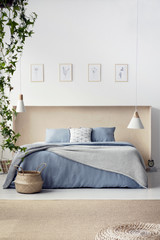 Real photo of king-size bed in wooden box standing in white room interior with wicker basket, two lamps, simple posters and carpet