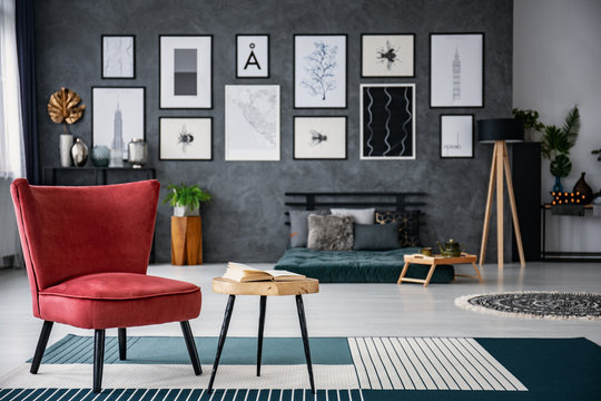 Red armchair next to table on carpet in dark grey living room interior with gallery in blurred background. Real photo