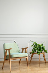 Real photo of an armchair standing next to a small table with a plant in simple room interior