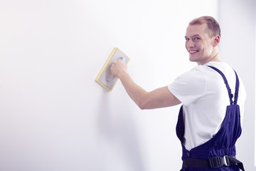 Young, happy wall painter in workwear posing, looking at the camera while smoothing down a white...