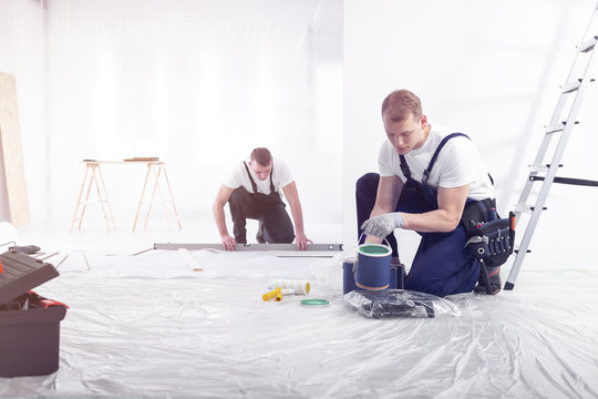 A man with a professional tool belt holding a can of green wall paint and a carpenter taking measurements in a white home renovation site interior