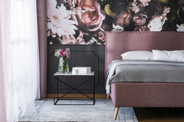 Flowers on table next to pink bed with grey sheets in bedroom interior with wallpaper. Real photo