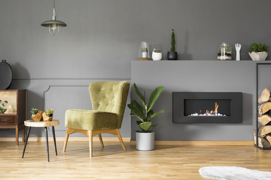 Lamp above green armchair and table in grey apartment interior with plant and fireplace. Real photo