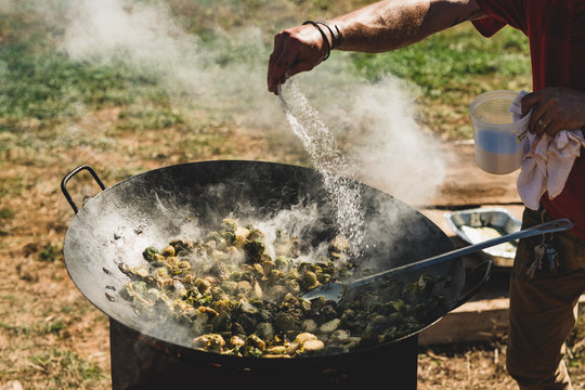 A man salts and cooks a wok of Brussels sprouts over an open fire