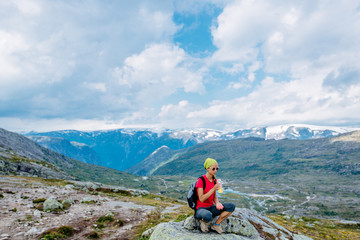 Fototapeta premium Travel in Norway, woman hiker in bright sportswear with backpack sitting on big stone and drinking water over scandinavian summer view.