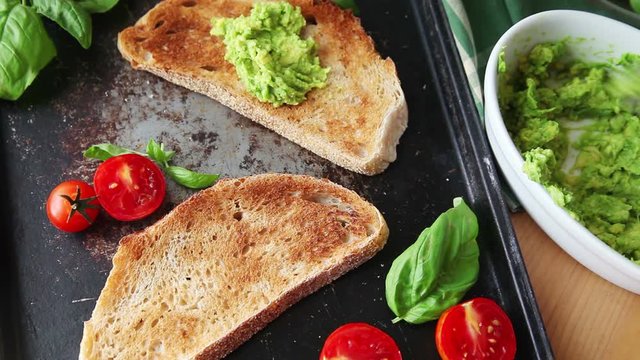 A woman spreads mashed avocado on toast, then adds cherry tomatoes 