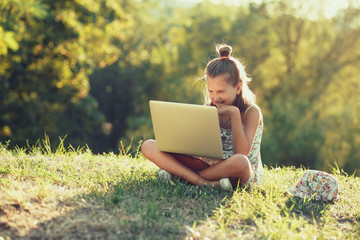 little girl is talking on a laptop while sitting on the grass in the sun. Dressed in a sarafan and...