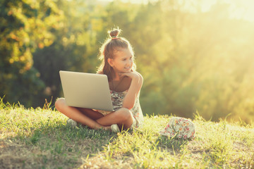 little girl is talking on a laptop while sitting on the grass in the sun. Dressed in a sarafan and...