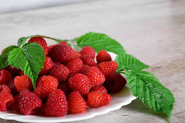  Red fresh raspberries on white rustic wood background. Plate with natural ripe organic berries with green leaves.flat lay with copy space